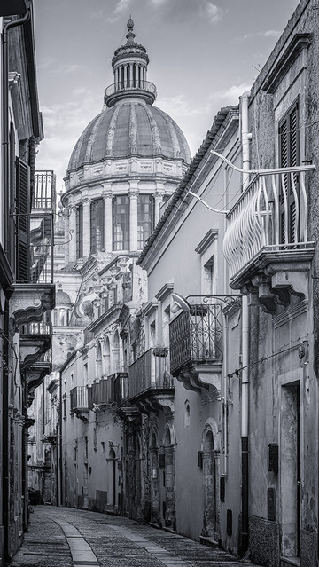 Black and white photography showing an historic lane lined with old buildings in Ragusa Sicily Italy. The dome of the Ragusa Cathedral is in the background. 