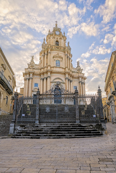 Church of San Giorgio in Ragusa Sicily. the main facade has three tiers divided by sculpted cornices and topped by a belfry. A single bell in an arch under a clock and a small crown shaped dome tops the structure. The church sits atop a flight of wide stone steps, while a detailed wrought iron gate encloses the property. 