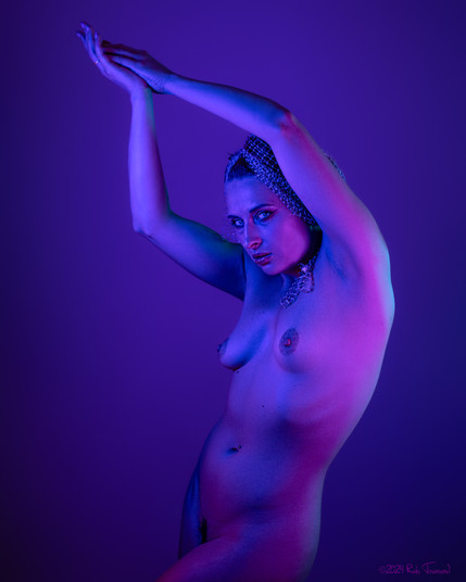 Art model Aurora poses nude in purple light with her arms outstretched over her head. She has a chain veil wrapping up her hair, and her piercing blue eyes are looking into the camera.