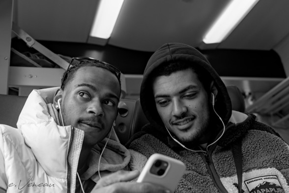 Two young people sitting on the train share a video on a cell phone. The one on the left glances out the window to check the name of the stop.