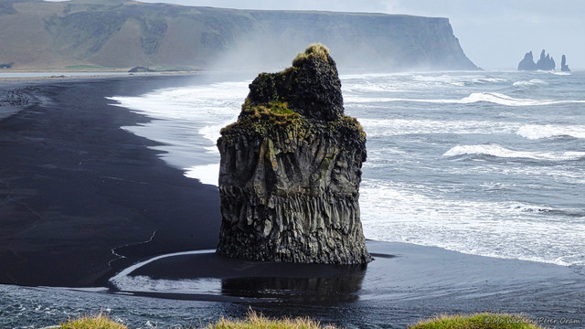 A photo of a black sand beach, stretching away from the viewpoint. The sea is on the right, sweeping stormy, white-capped waves onto the sands. In the foreground is a stream of water coming from the left to meet the sea and, on its bank, a twisted stump of columnar grey rock with vegetation on the horizontal tops. The black sand stretches away in a curve towards the right, where there is a cliff face. Off the end of this is a set of jagged sea stacks. Spray is rising from the sea, suggesting strong offshore winds.