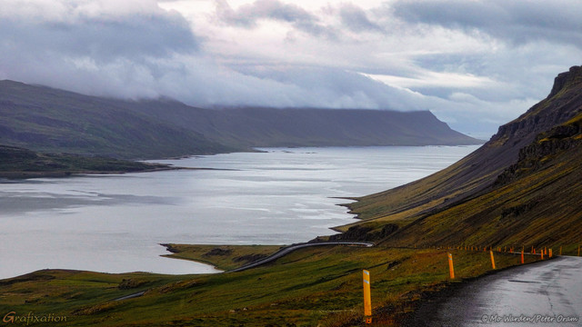 A photo of a landscape. A silver-grey fjord stretches from the bottom left to the centre right of the shot, its surface rippled by the wind. The sky is grey and cloudy, and a mountain on the opposite side of the water is cloud-capped. The viewpoint is from a roadside, high above the water surface. The rough tarmac road is to the right of the shot and drops steeply to hairpin bends lower down. There is no barrier at the road's edge, only snow poles. The scene is tranquil and peaceful.