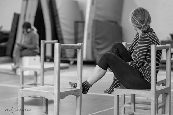 Sitting on a wooden chair, her feet in socks placed on another wooden chair, a woman turns her back to us. Both his sweater and his socks are striped in black and white.