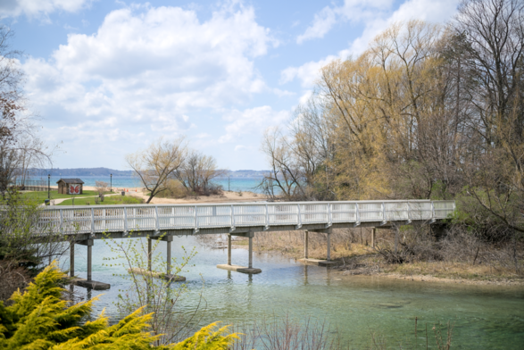 The photograph captures a serene landscape featuring a white wooden footbridge crossing a clear, shallow body of water. The bridge leads to a sandy beach area with sparse trees and a small building in the background. The water below the bridge is calm and transparent, reflecting the sky and surrounding vegetation. Beyond the bridge, the sandy beach transitions into a picturesque shoreline with trees, some of which are budding with spring growth. The horizon reveals a distant view of a large body of water, likely a lake, under a sky filled with fluffy white clouds.