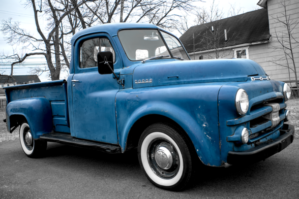 The photograph features a vintage blue Dodge pickup truck parked on a paved surface. The truck has a classic, rounded design typical of mid-20th century models, with a robust front grille, round headlights, and a slightly weathered appearance. The body of the truck is blue color, showing signs of age and use, such as minor scratches and faded paint. The white wall tires add to the nostalgic feel of the vehicle.