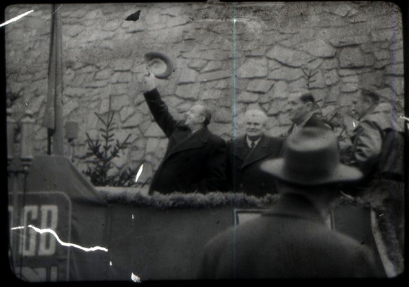 A black and white still of a group of men near a wall. One of them is waving his hat above his head.