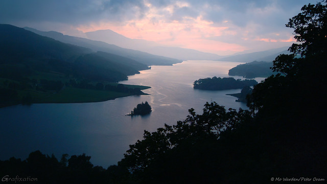 A photo of a valley, with a still loch running from lower left to top right of the shot. The distance is misty, and the low light has converted the mountain flanks into silhouettes. A line of dark trees is masking the view from the top right corner to the middle centre, and the water is lit more strongly in the distance by the setting sun, which isn't visible. The cloud is quite heavy but the light filtering through is soft pastel pink, the rest of the scene is in a cyan and blue palette. It's tranquil, and very calm.
