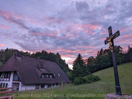 Pink and purple clouds with light yellow on horizon above a black forest house and a gold crucifix