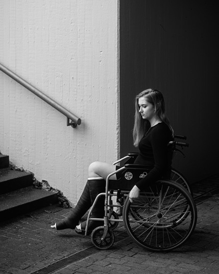 Junge Frau sitzt mit zwei Gipsbeinen in einem Rollstuhl am Fuß einer Treppe und schaut traurig zu Boden.