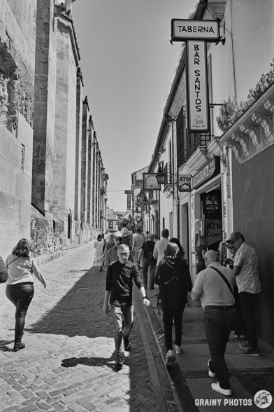 This is a black-and-white film photo of a narrow cobbled street with the Mezquita-Catedral de Córdoba wall on the left and shops and cafes on the right. The street is busy with tourists.