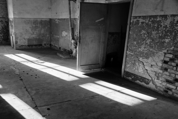 Innenansicht eines alten, leeren Gebäudes. Sonne fällt flach in den Raum. Auf der rechten Seite sieht man eine geöffnete Tür. Die Wände haben zum Teil keinen Putz mehr. 