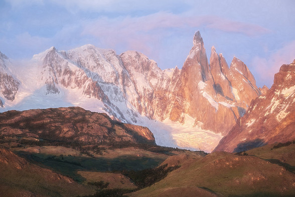 Sunrise in the Patagonia area of Argentina brings a burst of color on the snow-capped mountain peaks. Within minutes, the dark brown/maroon-colored rocks become orange and red while the clouds above the rugged peaks glow pink. Green valley in the foreground contrasts with the jagged peaks and vibrant sky. The long early day shadows provide texture to the foreground valleys. 