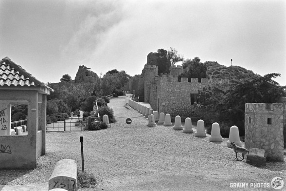 A black-and-white film photo of the entrance road to the Baterías de Castillitos. In the foreground is a checkpoint hut, and the road then forks as it continues into the complex. The buildings in the complex were built to imitate a medieval castle - hence the name Castillitos. Some of the buildings can be seen on the RHS.