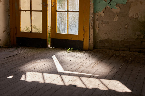 Eine gelbe Pendeltür in einem verlassenen Gebäude. Durch einen Türspalt fällt Sonnenlicht in den Raum. 