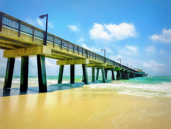 A long, sturdy pier extends over clear turquoise waters, with waves gently crashing against its black pillars. The sandy beach glows under a bright blue sky dotted with fluffy white clouds. Photo Art by Debra Martz