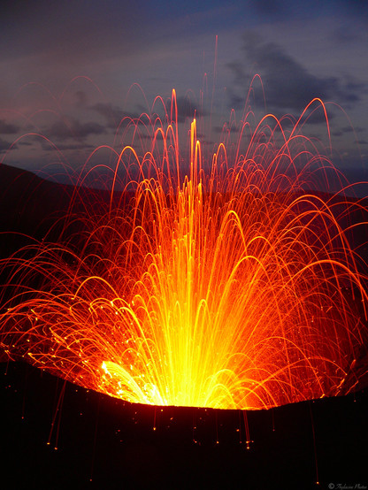 A vertical zoom in of a volcano vent, taken with a slow 8 second exposure to capture the motion.  It's spurting lava into the early evening sky in a shower of red and yellow parabolic arcs, emanating from the centre and falling all around the vent.