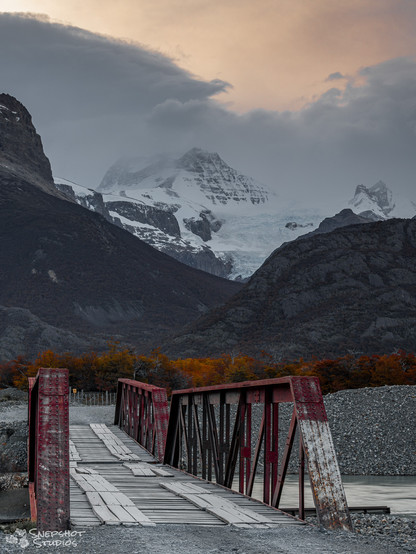 A wobbly looking one lane steel bridge with a patched up wooden deck across a river. In the background, an autumn forest, high mountains and a glacier