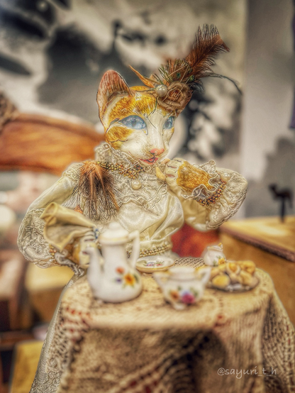 A cat shaped antique music box. A cat is dressed in antique dress and hat, and there’s a tea set in front