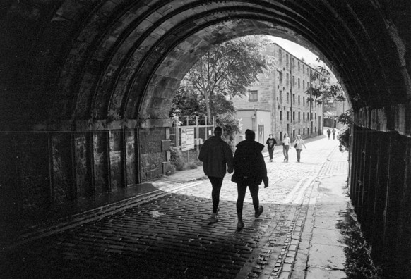 Black and white mostly low key image taken from under an arched railway bridge, showing people walking on a stone-paved street. Stone tenement building are visible on the distant left. Two silhouetted figures take the centre of the frame, one in med-step with foot poised for ever in the air!