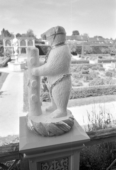 Bear with ragged staff, out of focus fountain in the left hand background. Black and white portrait format photo. SMC Pentax M 28mm f/3.5.