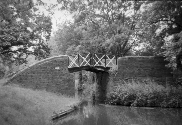 Black and white photo of a canal bridge with brick abutments and black and white beams and crossed guard rails. The two parts of the bridge have a small gap for the rope. The bridge is number 52. Out of focus trees above, banks and the still water in the foreground. A peaceful scene.