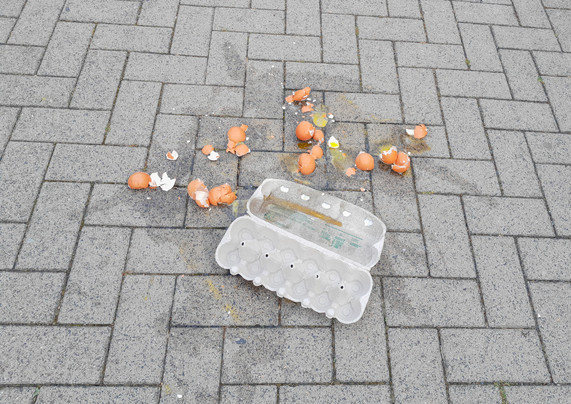 Eine leere Eierverpackung auf einem gepflasterten Weg, dahinter viele Eierschalen