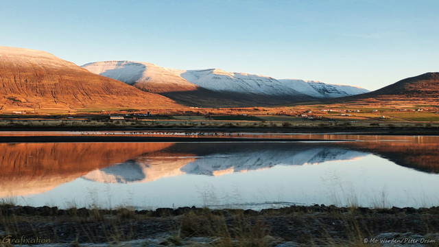 A photo of a landscape under a clear cyan sky. The foreground is a river bank, the still waters beyond reflecting the scene on the opposite bank. On the other side of the water is a dark sand bank before the shoreline, between these two in a protected area of river are dozens of water-birds. The distant horizon is a line of snow-capped mountains, nestled in a valley between them are multiple farm buildings and homes. The lowering sun is casting a softly orange-tinted light from the right side.