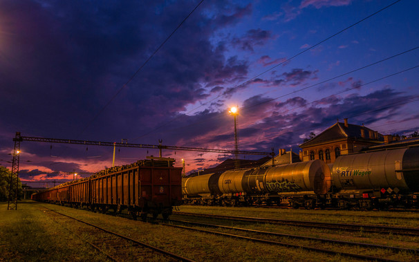 Güterbahnhof, Abstellgleise während der blauen Stunde. Eine Laterne, Stromleitungen, Gleise, Waggons.
Im Hintergrund das Bahnhofsgebäude