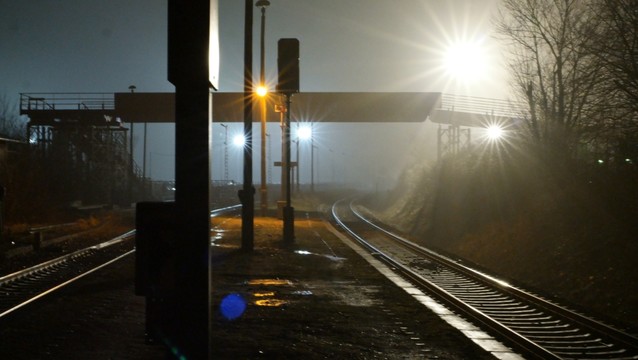 Ein unbefestigter Bahnsteig zwischen zwei Gleisen. Darauf sind mehrere Masten mit verschiedenen dunklen Aufbauen. Dahinter eine Fußgängerbrücke und mehrere Lichter, die bläulich, weiß und gelblich leuchten, und Blendenstrahlen werfen. Die Lichter spiegeln sich in Pfützen und in den Schienen.