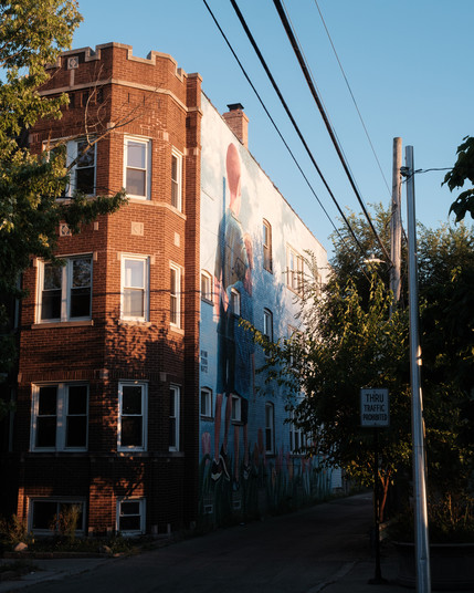 A mural of two children against a sky blue backdrop on the side of a brick building in an alleyway.