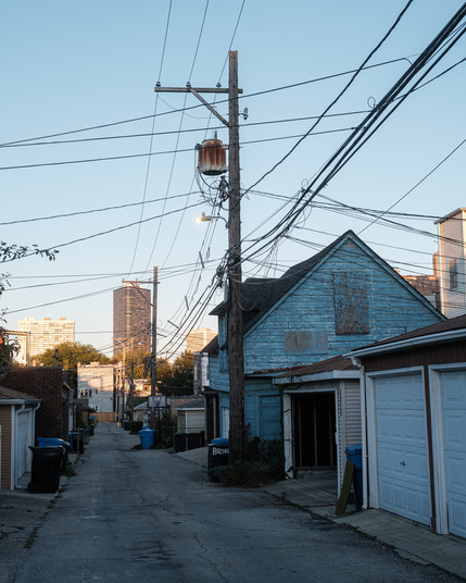 An old blue garage with faded paint and sagging boards sits in juxtaposition to the modern high rise in the distance. A criss-cross of powerlines drapes over the composition.