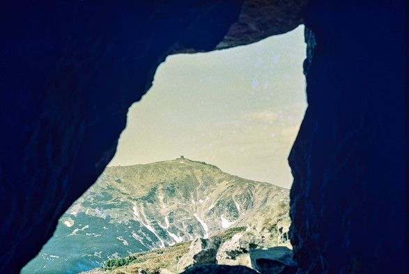 The image shows a scenic view of a mountain, framed by the dark edges of what appears to be the entrance of a cave. The vantage point from the cave looks out onto a distant mountain, with rocky and grassy slopes, some of which are still covered in patches of snow. On the peak of the mountain stands a small structure, possibly a weather station or an observatory. The lighting suggests daylight, with clear skies and a soft glow. The overall effect creates a sense of looking out from a hidden or sheltered spot towards a vast, natural landscape.