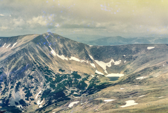 The image captures a panoramic view of a mountainous landscape, showcasing a large, rugged mountain range. The slopes are a mix of brown, rocky terrain with patches of snow still clinging to the surface, even in the warmer seasons. Dark green vegetation dots the mountainside, contrasting against the lighter brown tones. In the valley below, there is a small, serene lake surrounded by more snow, nestled between the mountains. The sky above is cloudy, casting a soft, diffused light over the scene, creating a moody, atmospheric feeling. In the distance, faint outlines of additional mountain ranges are visible, fading into the misty horizon.