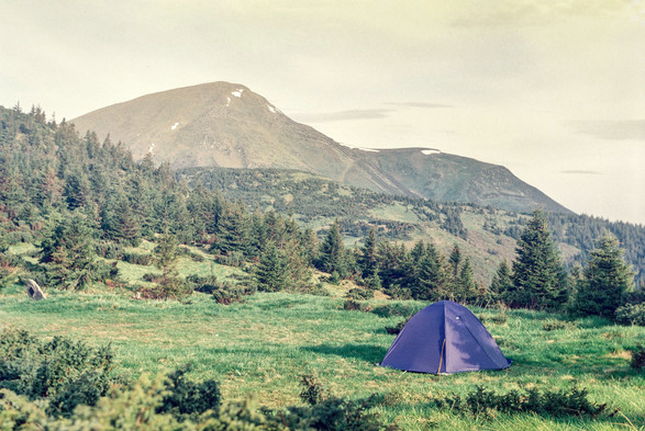 The image shows a scenic mountain landscape with a camping tent in the foreground. Here's a detailed description in English:

In the foreground, there's a small purple tent pitched on a grassy meadow. The meadow is lush and green, dotted with low shrubs and wildflowers.

Behind the tent, the landscape rises into forested hills. These hills are covered with dense evergreen trees, primarily pine or fir. The forest extends up the slopes, creating a dark green blanket over the lower elevations.

In the background, a prominent mountain peak dominates the skyline. This mountain appears to be quite tall, with a distinct triangular shape. The upper portions of the mountain are bare rock, with some patches of snow still visible near the summit, suggesting it's a high-altitude peak.

The sky is pale and hazy, giving the scene a soft, ethereal quality. This muted atmosphere lends a sense of tranquility to the entire landscape.

Overall, the image captures the essence of wilderness camping, with the small tent dwarfed by the grand scale of the surrounding nature. It evokes feelings of solitude, adventure, and connection with the natural world.