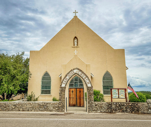 A historic church building stands with a beige facade and arched windows, featuring a small statue in a niche above the entrance. The church is named "San Jose Church" and includes a wooden gate and a stone wall in front. Photography by Debra Martz