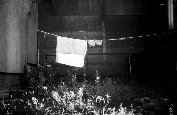 The black-and-white image shows an outdoor scene with a clothesline stretched between two posts. On the line, there is a large towel or sheet hanging in the center, and smaller items, like pieces of clothing or rags, hanging to the right. The background includes dark walls or a fence, creating a stark contrast with the lighter objects in the foreground. Vegetation, including tall grass and weeds, fills the lower part of the image, with sunlight filtering through the plants and casting shadows. The overall mood of the image feels quiet and somewhat melancholic due to its monochrome tone and the empty, abandoned atmosphere.