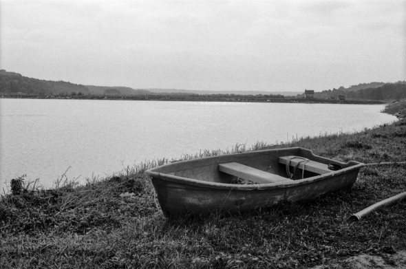The image depicts a serene, grayscale scene of a small boat resting on the shore of a large body of water. The boat appears weathered and is positioned on the grass near the edge of the water. In the background, the water stretches across the frame, with distant hills and some trees visible on the horizon. A few small buildings can be seen far in the distance, adding to the sense of isolation and quiet. The overcast sky and overall gray tones create a calm, somewhat melancholic atmosphere.