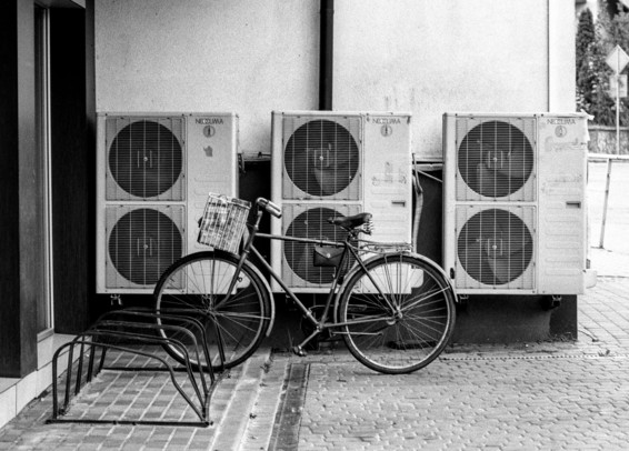 The image is a black and white photograph depicting a bicycle parked in front of a row of air conditioning units. The bike has a basket attached to its handlebars and is resting against the AC units, which are lined up on the wall. The setting appears to be an urban area, with the bicycle placed next to an empty bike rack on a paved sidewalk. The scene is simple and industrial, featuring a contrast between the vintage-looking bike and the modern air conditioning units, labeled "NEOCLIMA."