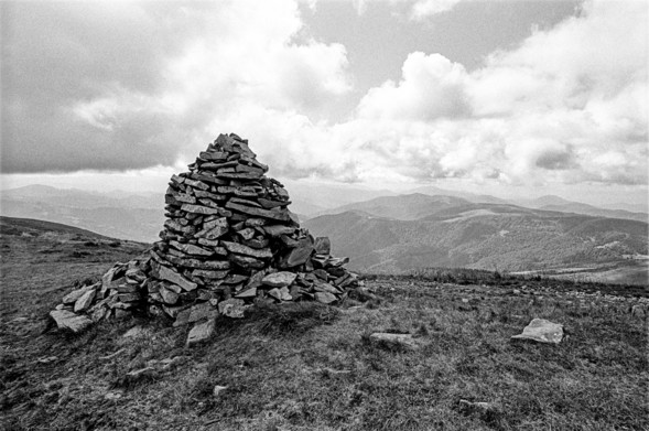 The image shows a stone cairn, a stack of rocks carefully arranged into a conical shape, situated on a grassy hillside. It appears to be part of a remote, mountainous landscape, with rolling hills and forested areas in the distance. The sky is filled with clouds, adding a dramatic backdrop to the scene. The photo is black and white, giving it a timeless and serene quality, emphasizing the textures of the rocks and the vastness of the landscape beyond. The image evokes a sense of isolation and tranquility, with no signs of human activity apart from the cairn itself.