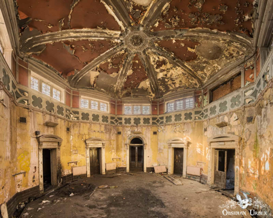 old decaying room, a rotunda with yellow walls an multiple doors. The roof is domed and trussed, with red peeling paint