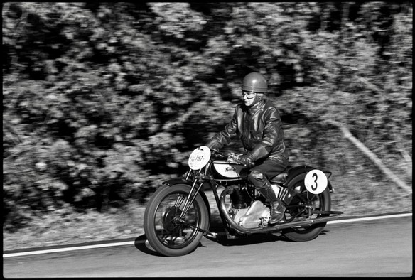 Black and white film photography. A vintage Norton CS1 motorcycle with a rider dressed in a leather jacket rides around a forest