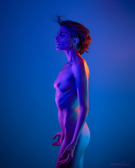 A photo of art model Roarie Yum by photographer Rab Townsend. Roarie is posing nude in blue, purple, and orange light against a blue background. Roarie is standing in profile, facing the left of frame, with hands dropped at their side. The orange light is cast against their back and shoulders. Roarie's eyes are closed, and their hair is caught in motion.