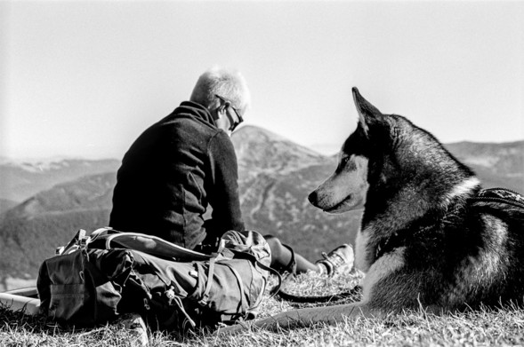 The black-and-white image shows a serene moment in nature, featuring a woman sitting on the ground, viewed from behind, with a dog resting beside her. The woman has short light-colored hair, wears a dark jacket, and seems to be gazing toward a mountain landscape in the distance. Their backpack lies next to her, suggesting she might be on a hiking trip. The husky dog is lying down, also looking in the same direction. The backdrop includes rolling mountain hills, creating a peaceful and contemplative scene.