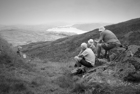 Black and white photo, three people wearing white hats sitting on a hill, looking towards Windermere in the distance. Another person looks towards them.