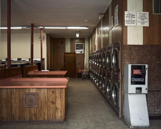 This image shows the interior of a laundromat. The space is filled with washing machines arranged along the wall, and there are wooden tables with pink countertops in the foreground. The walls have a warm, wooden texture, creating a cozy atmosphere. You can hear the faint sounds of machines running and the occasional rustling of laundry. There's a change machine on one side, and a clock on the wall indicating the time.