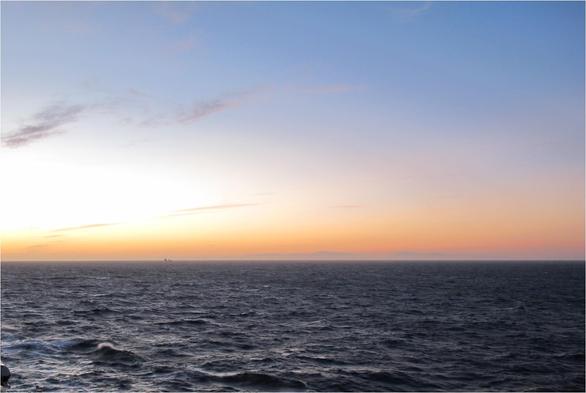 At sea. A ship is on the horizon. It is around sunset.