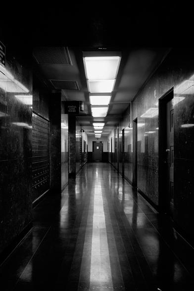The image shows a long, narrow hallway in a building. The floor is shiny and dark, reflecting light from overhead fluorescent lights. On one side, there are rows of postal lock boxes, and there are doors on both sides of the hallway. The walls are dark and glossy, creating an atmosphere that feels both modern and a bit somber.