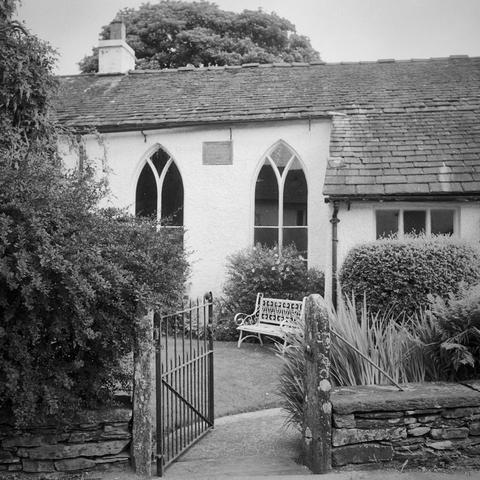 An open gate leads into a small garden with an iron seat in front of a modest chapel, part of a terrace of buildings, with beautiful pointed windows. There is a small plaque with indistinguishable writing. Black and white square photo (cropped).