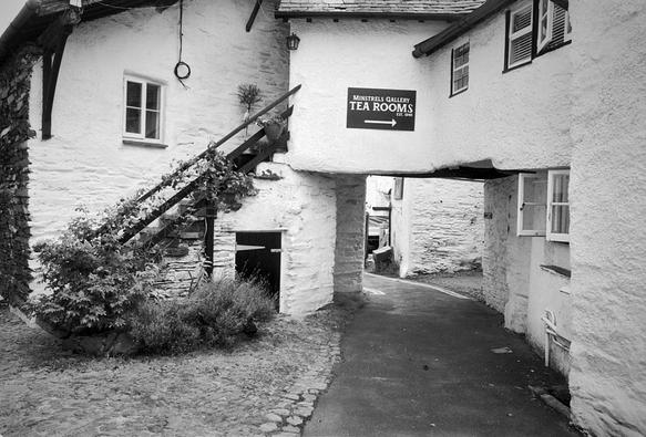 This black and white photo is of a rustic-looking white-rendered building, built like a bridge across the track coming from the bottom of the frame. Above the track is a sign that reads "Minstrels Gallery Tea Rooms Est 1946" with an arrow pointing to the right. To the left s a stair up to the first floor, with wooden banisters with climbing plants winding through them. Above the stair a wire hangs down, ending in a coil. Ahead under the "bridge" the track bends left, but it seems there is another track to the right.