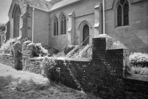 A stone-built church building from close by. The windows are stone-framed and pointed. There is a stone wall in front, with a gap for a gate; inside the wall, steps rise to a doorway in the church wall. Black and white photo.
